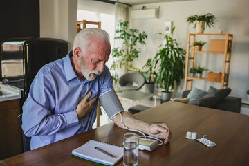 Senior man checking his blood pressure at home, pressing his chest while monitoring readings, with...