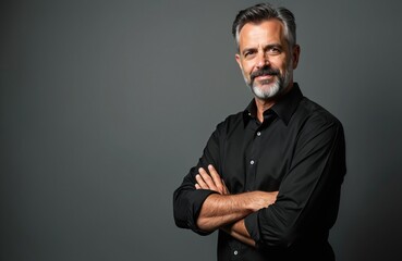 Fototapeta premium Portrait of confident middle aged man with grey beard, mustache smiles at camera. Wears black shirt, crosses arms on chest. Stylish professional businessman stands alone on grey studio background,