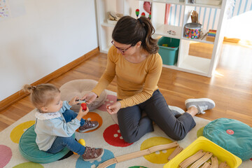 Caregiver and young girl sitting on a colorful rug, playing with wooden toys together in a bright, cozy playroom