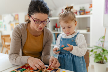 Smiling kindergarten teacher and toddler painting and crafting together at a bright classroom table, showing early learning, creativity, and child development in a warm preschool environment