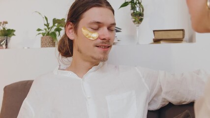 Man receives skincare treatment at home while a woman applies eye patches during self-care session