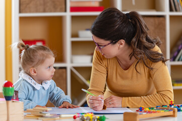Fototapeta premium Adult woman guiding a toddler during a drawing activity at a table with colorful toys, supporting early learning and creativity in a bright home or classroom setting