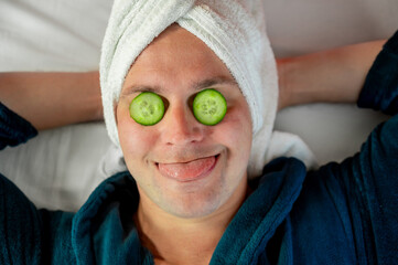 Man with cucumber eye mask in bathrobe, having fun during skincare