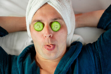 Man with cucumber eye mask in bathrobe, having fun during skincare