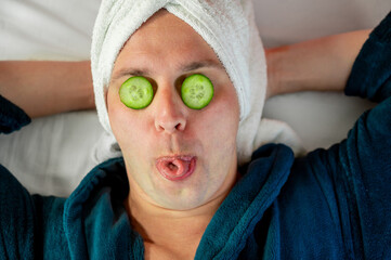 Man with cucumber eye mask in bathrobe, having fun during skincare