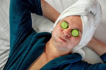 Man with cucumber eye mask in bathrobe, having fun during skincare