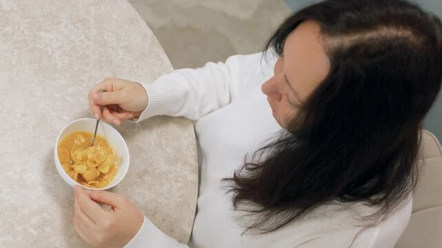 High angle view of woman in white sweater holding spoonful of corn flakes cereal over white bowl at round beige marble table, lifting spoon to mouth.