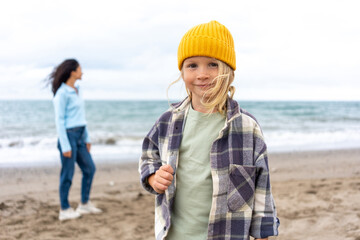 Smiling blond child in yellow beanie and plaid jacket standing on the beach, with his mother in a...