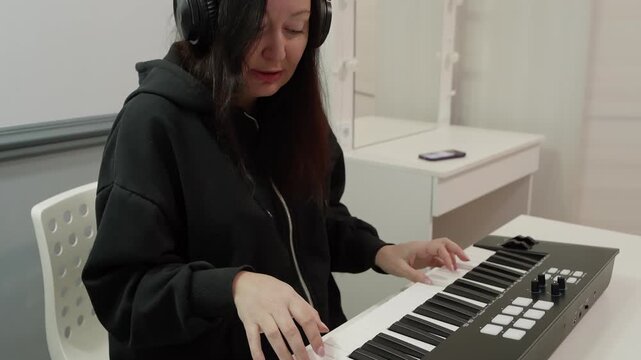 Asian woman in black hoodie and headphones sits on white chair playing MIDI keyboard synthesizer at white desk near window with curtains, expressing joy while creating music.