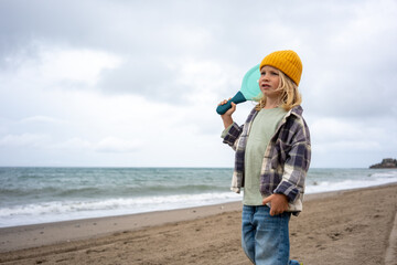 Blond boy in yellow beanie holding turquoise paddle on shoulder while standing on sandy beach near ocean waves, dressed in plaid jacket and jeans, looking thoughtful under cloudy sky