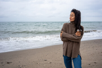Woman in brown turtleneck sweater holding coffee cup and crossing arms, standing on sandy beach near ocean waves, looking to the side with calm expression under cloudy sky