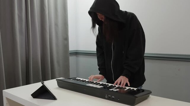Musician with long hair wearing black hooded sweatshirt with hood down stands playing MIDI keyboard controller on white desk in modern room with gray and white walls.