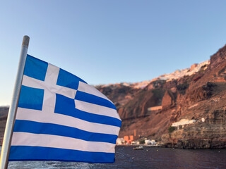 Greek flag waving proudly on a boat sailing near the island cliffside houses of Santorini, Aegean sea, basking in the glow of a warm sunset light