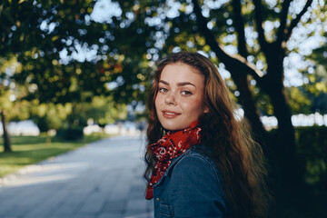 Fototapeta premium smiling woman with freckles and curly hair wearing denim jacket and red scarf outdoors in sunny park with green trees background