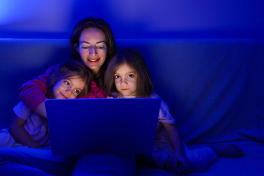 Happy Mother And Two Little Daughters Watching Movie On Laptop At Night Together
