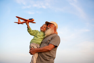 Joyful grandfather holding his happy blond grandson who is lifting a toy airplane into the sky, both enjoying playful time together under a clear blue sky.
