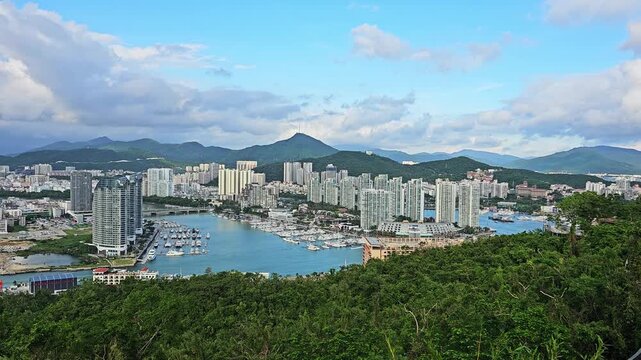 Scenic panoramic view of Sanya from Deer Turned Head Park, featuring a modern skyline, marina, mountains, lush greenery, and waterfront under a bright tropical sky.