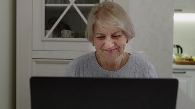 Smiling Senior woman in gray cable-knit sweater working on laptop from side view. Focused typing showing productivity, digital work and technology proficiency in bright home office setting.
