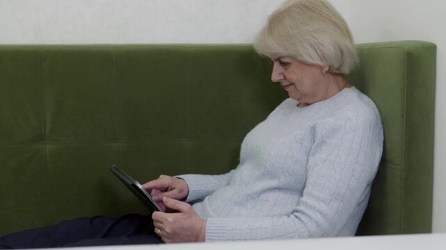 Elderly woman in gray cable-knit sweater working on tablet at table with engaged expression. Side view showing productivity, digital work and technology proficiency in bright home setting.