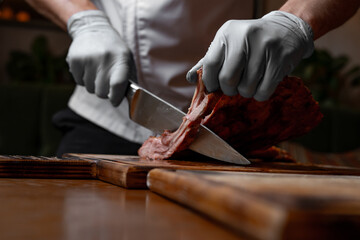 Chef cutting juicy grilled pork ribs on wooden board, close up of hands with knife slicing roasted barbecue meat in restaurant kitchen