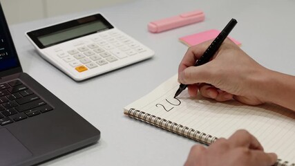 Close-up of hand writing 2026 in notebook with pen on desk with calculator and laptop. Concept of planning, budgeting, financial goals, new year strategy, productivity and personal development.