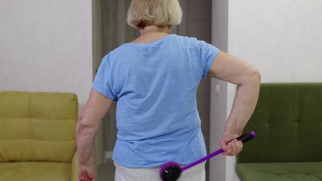 Elderly woman in blue shirt performing self massage on lower back with purple handled massager brush. Therapeutic wellness routine for muscle tension and back pain relief at home.