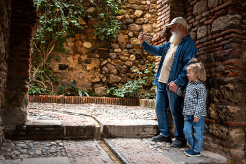 Fototapeta premium An elderly man and a young child taking a selfie together in a stone-walled garden with plants in the background