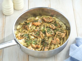 A pan filled with cooked salmon and sliced fennel, seasoned with herbs, sits on a weathered wooden table. Soft, natural light illuminates the scene