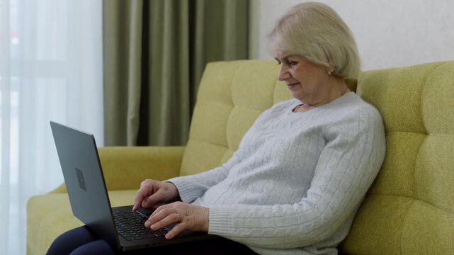 Elderly woman in light gray sweater and black pants working on laptop with genuine happy expression. Positive emotion showing enjoyable online content and technology satisfaction on yellow couch.