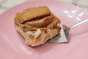 Freshly baked cinnamon roll topped with creamy biscoff spread and a lotus cookie, being served on a metal spatula on a glossy pink plate