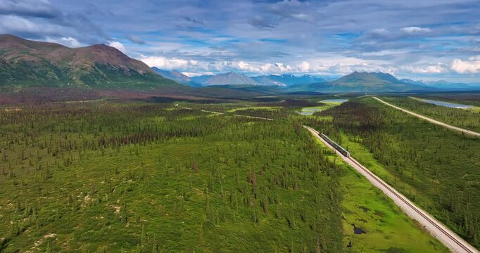 Train Journey Through Vast Alaskan Tundra and Boreal Forest. An aerial perspective captures a modern passenger train traversing a railway line through immense green tundra
