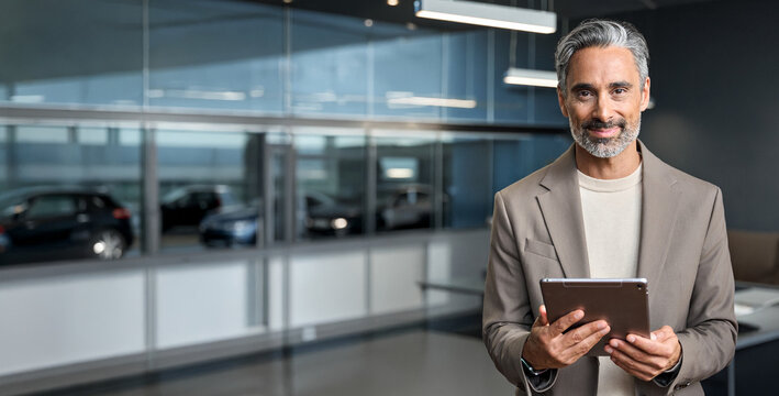 Happy male manager auto dealer salesman standing in car dealership office with tablet looking at camera. Car leasing concept. Buying new car, vehicle trade and travel car hire. Banner