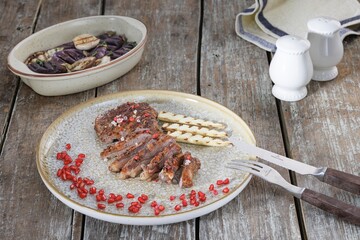 A perfectly cooked and artfully sliced steak is presented on a plate with grilled vegetables, adorned with bright red pomegranate seeds and served with a side of eggplant