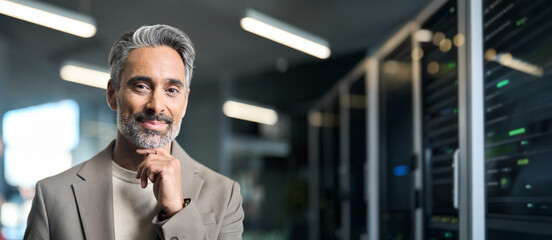 Happy mature man engineer wearing suit in data center work office. Middle aged manager looking at camera standing in server room security warehouse background. Banner, digitalization concept.