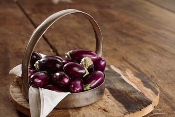Petite, purple eggplants fill a metal basket, nestled on a linen cloth. They sit on a rough-hewn wooden board atop a rustic table during daylight hours