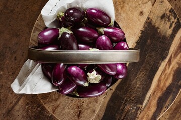 A multitude of miniature purple eggplants are piled high in a metal container lined with fabric, set against a backdrop of a rustic wooden surface