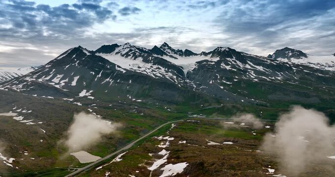 Foggy Road and Snow-Capped Peaks in Alaskan Mountain Pass. An elevated view captures a winding road and patches of fog in an Alaskan mountain pass