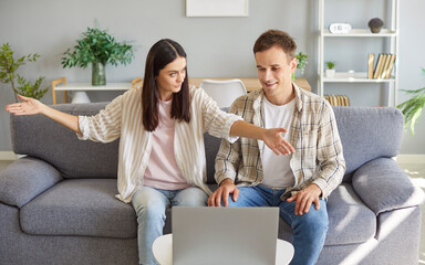 Young cheerful smiling couple man and woman sitting on sofa in living room at home using laptop together discussing repair projects planning renovation or choosing furniture for new flat