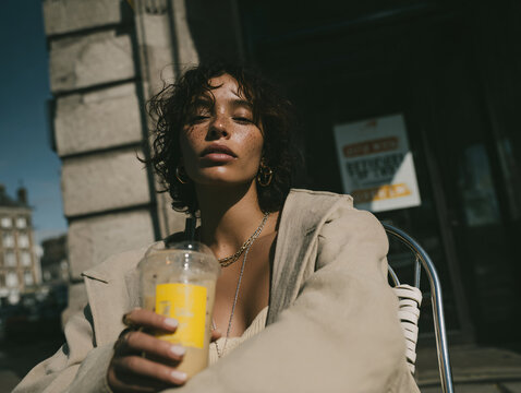 a person sitting outdoors, holding a beverage in a clear cup with a straw. They are wearing a black tank top and layered gold necklaces. The background features a stone building with a window