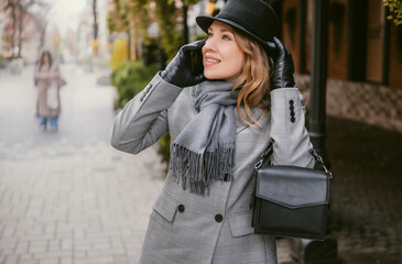 Fototapeta premium Blonde business woman talking on smartphone on autumn city street on cloudy cool day in gray suit scarf black hat and gloves showing focused confident stylish urban mood walking to important meeting