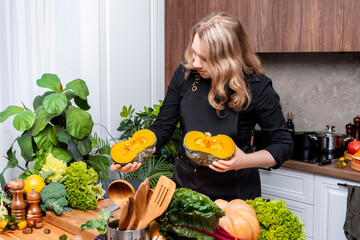 Smiling young woman in black apron holding large green pumpkin on wooden kitchen table with fresh...