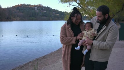 Cheerful lakeside walk shows smiling parents carrying their newborn baby beside calm water, surrounded by bright fall trees and a peaceful shoreline atmosphere with soft daylight.