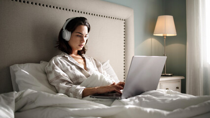 Young woman wearing headphones is sitting in bed with a laptop, surrounded by soft pillows and...