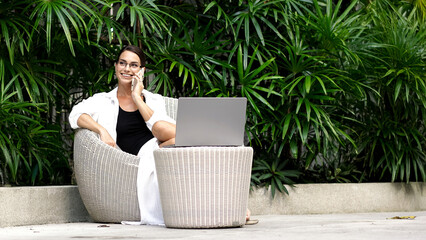 Young woman in casual attire sitting on a comfortable chair, using a laptop outdoors surrounded by lush greenery, enjoying a relaxed moment of remote work and connection © wedmoments.stock