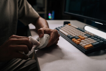Close-up of man disinfecting computer mouse with white wet wipe, ensuring cleanliness and hygiene on desktop accessories near keyboard, promoting health and maintenance for office equipment.