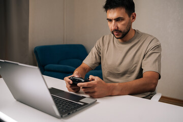 Portrait of young man concentrating playing, controlling computer game using wireless gamepad controller sitting at desk with laptop, enjoying leisure time and digital entertainment at home.