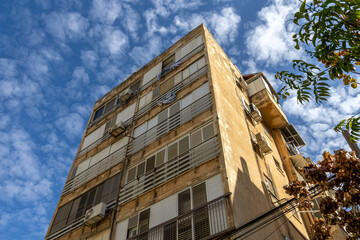 Weathered Israeli residential building with balconies laundry shutters and external AC units under blue sky. Aged stucco facade and urban neighborhood housing