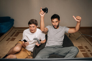Happy excited two friends sitting on sofa cheer and raise controllers in celebration as they play video games together at home, smiling and enjoying competitive, casual gaming night.