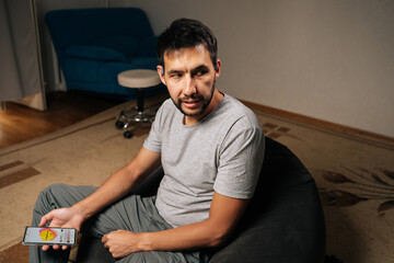 Bearded man wearing bone conduction headphones relaxes at home, focused on smartphone while streaming music and podcasts for private, comfortable listening experience.