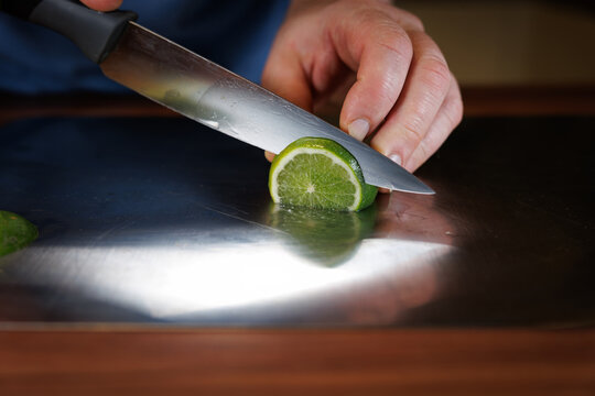 Close-up of a person slicing a fresh lime with a sharp kitchen knife on a stainless-steel cutting board